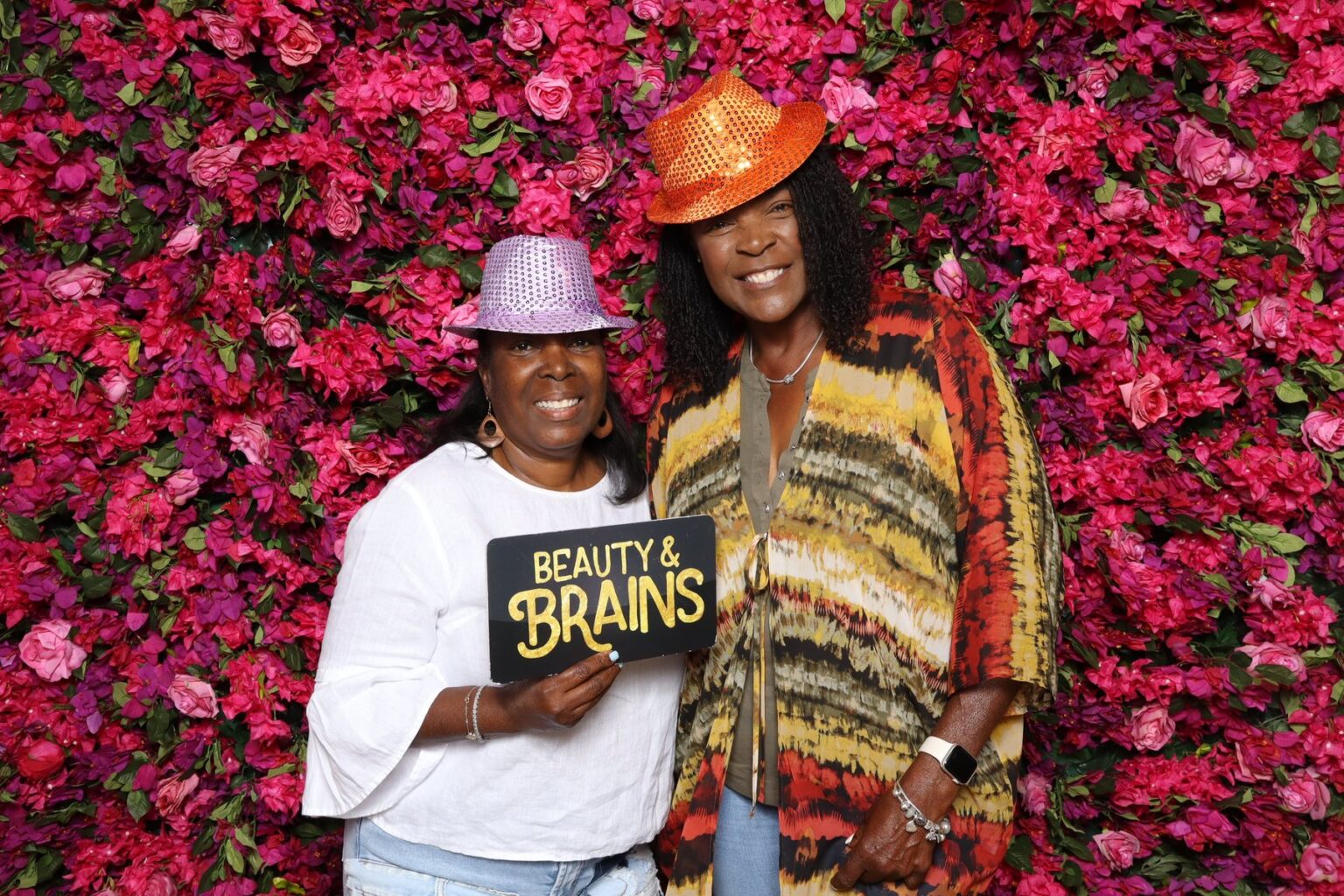 Two guests holding Beauty and Brains props at a pink flower wall photo booth