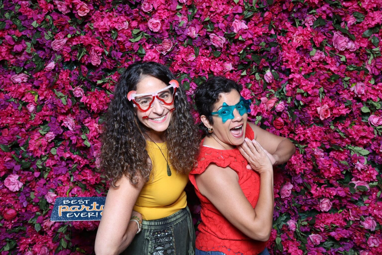 Two guests in prop glasses laughing at a pink flower wall photo booth