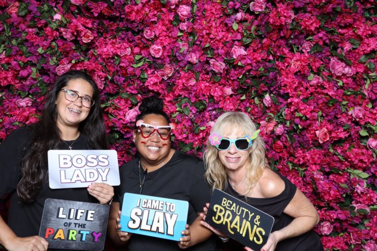 Three guests holding props at a pink flower wall photo booth