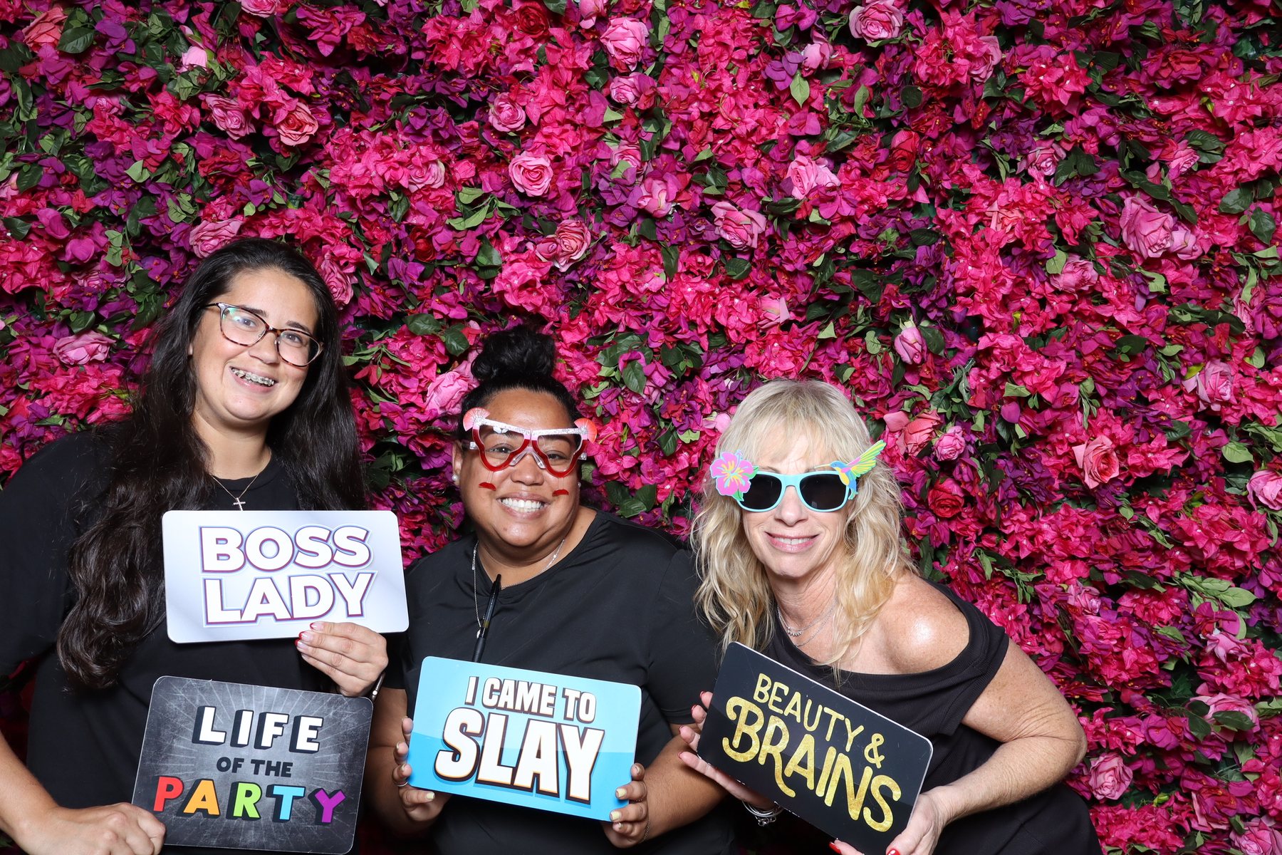 Three guests holding props at a pink flower wall photo booth